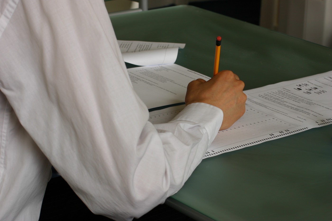 Una mano escribiendo con lápiz sobre un examen en papel, sola en un pupitre.
